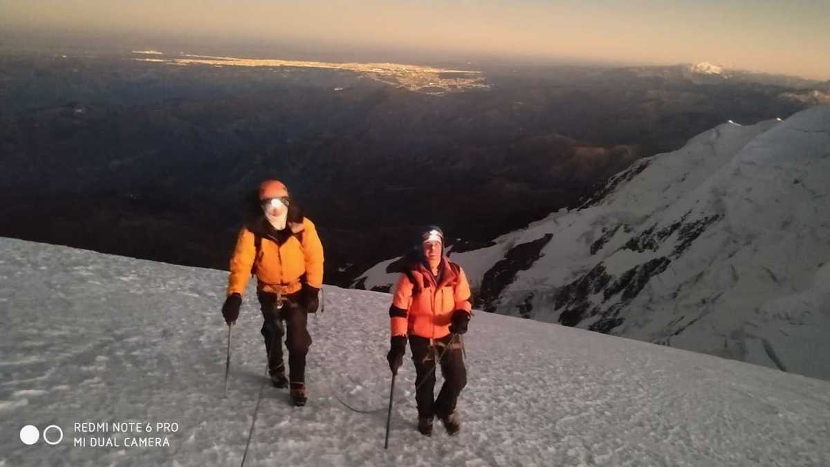 Two men approach the summit of Illimani at sunrise. The lights of La Paz can be seen in the background.
