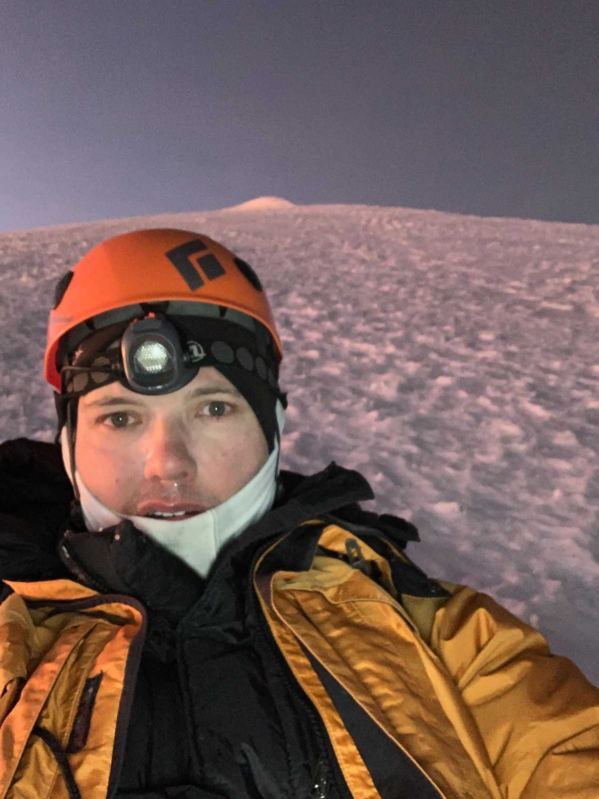 A climber looks very cold and tired. The summit of Illimani is still far above him.